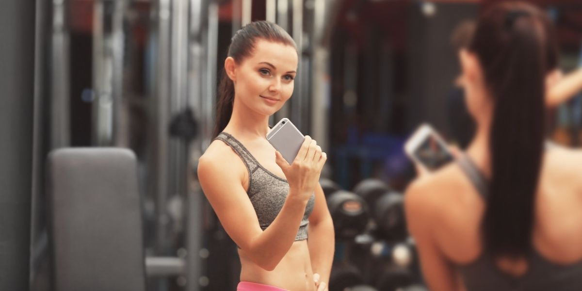 ジムでスマートフォンを使いながら筋トレをする女性の姿。筋トレアプリや動画を活用してトレーニングを継続する様子。