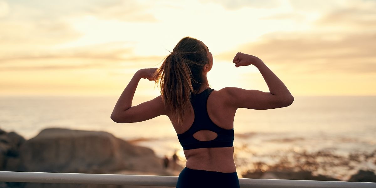 筋トレを習慣化し、海辺の夕日を背にして腕の筋肉をアピールする女性の後ろ姿。卒業後も続ける筋トレ習慣の大切さを伝えるイメージ写真。
