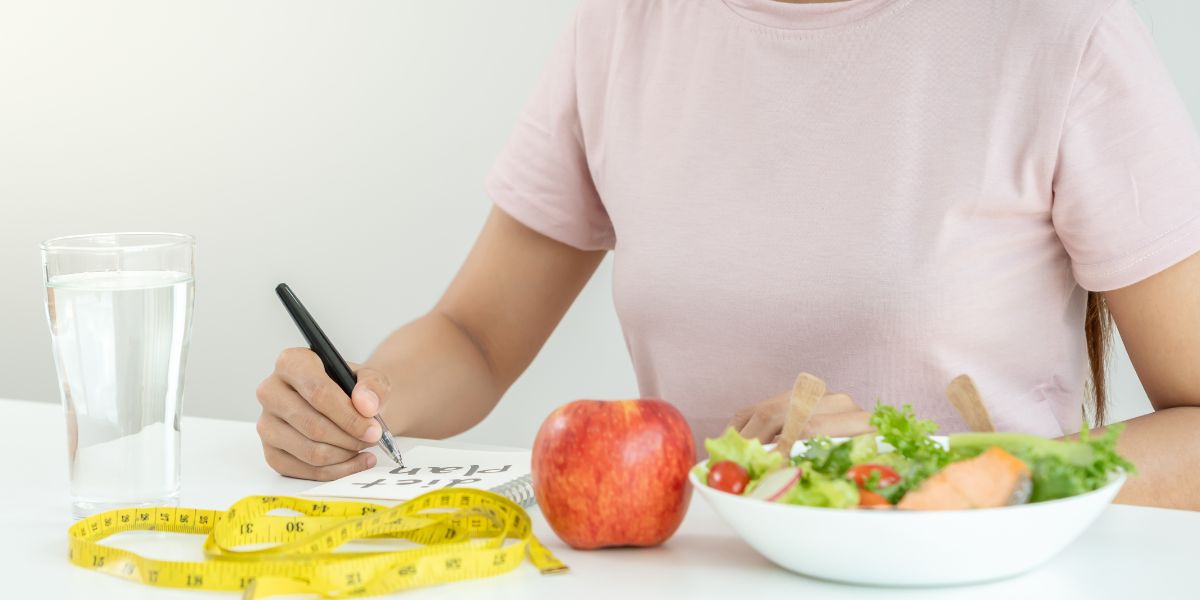 食事とトレーニング計画をノートに書き込む女性の手元。水・リンゴ・メジャー・サラダが並び、PFC設計と継続のイメージ。
